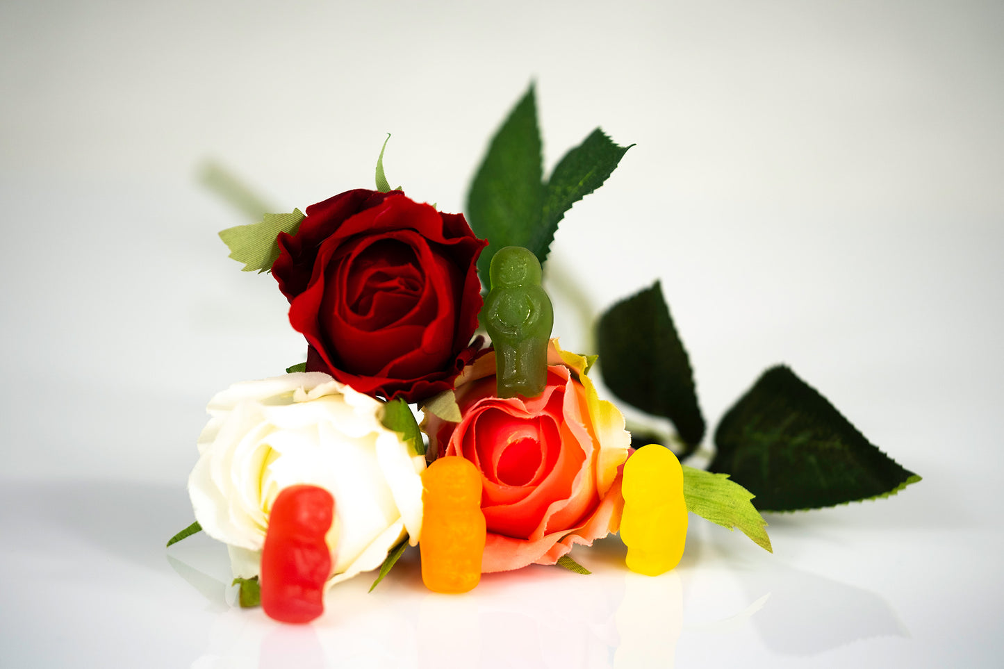 Colourful gummy figures inhabiting a miniature arrangement of artificial roses and green leaves on a reflective white surface.