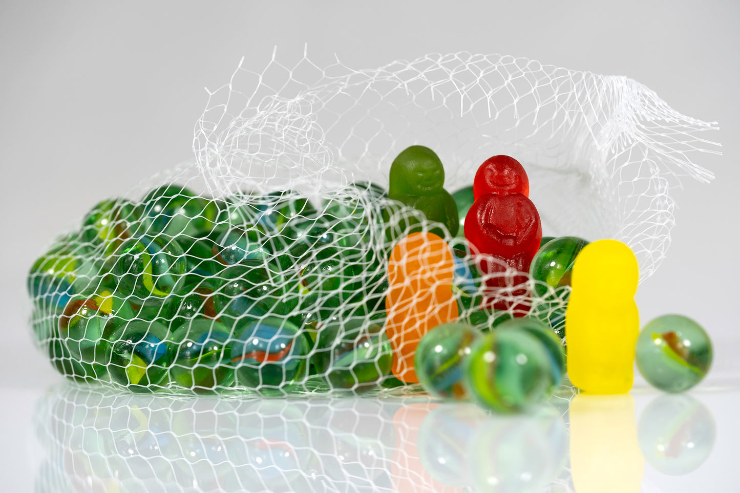 Green glass marbles spilling from an open mesh bag surrounded by colourful gummy figures and reflected on a white surface.