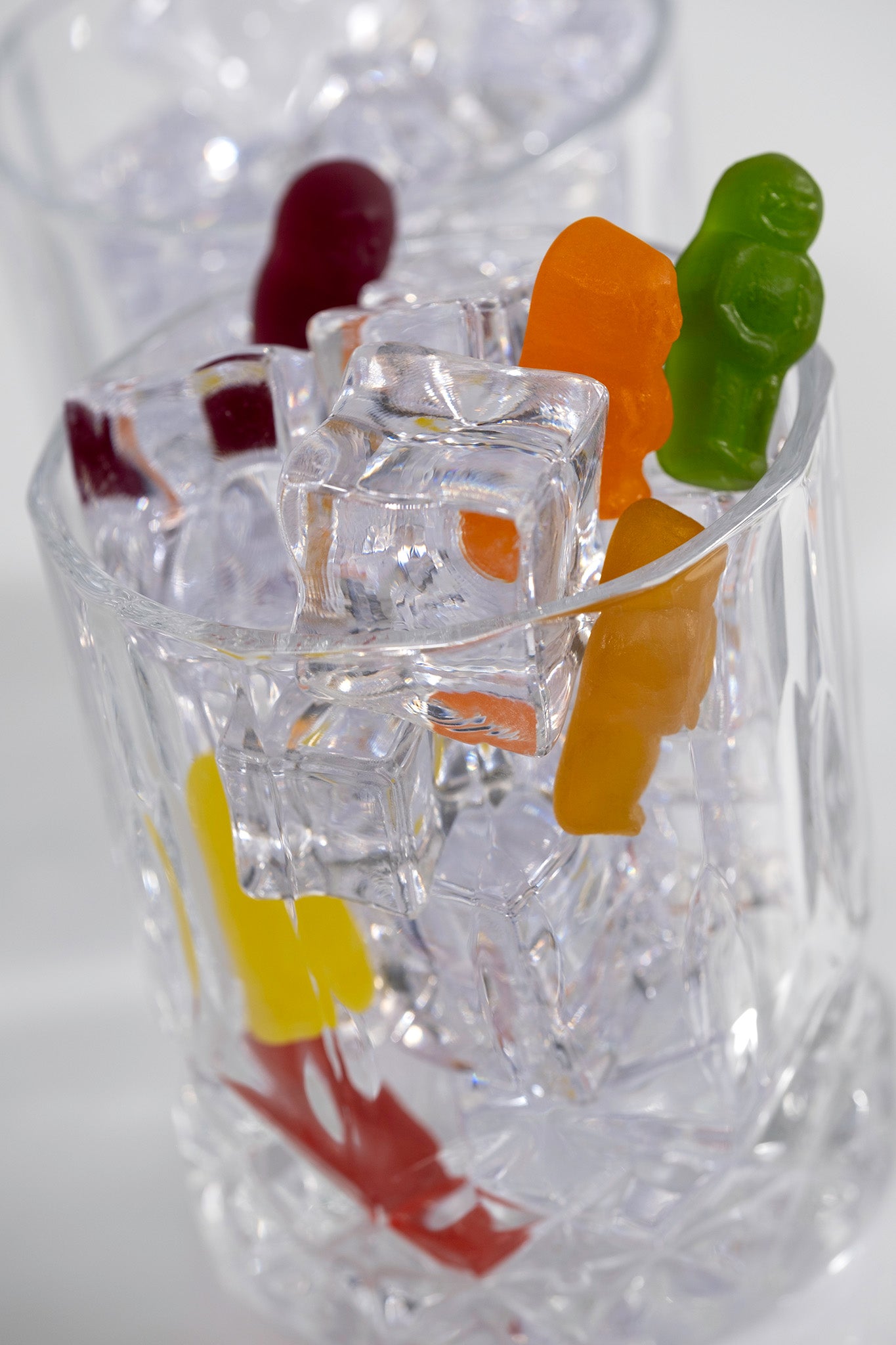 Portrait still life of colourful gummy figures surrounded by transparent ice cubes and glass, with refracted red and yellow forms visible below.