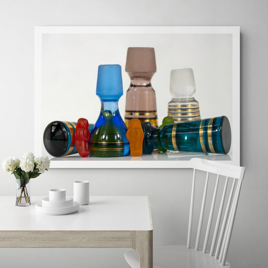 Large photographic artwork featuring colourful gummy figures among vintage blue, pink and clear glass stoppers, displayed above a contemporary dining table.