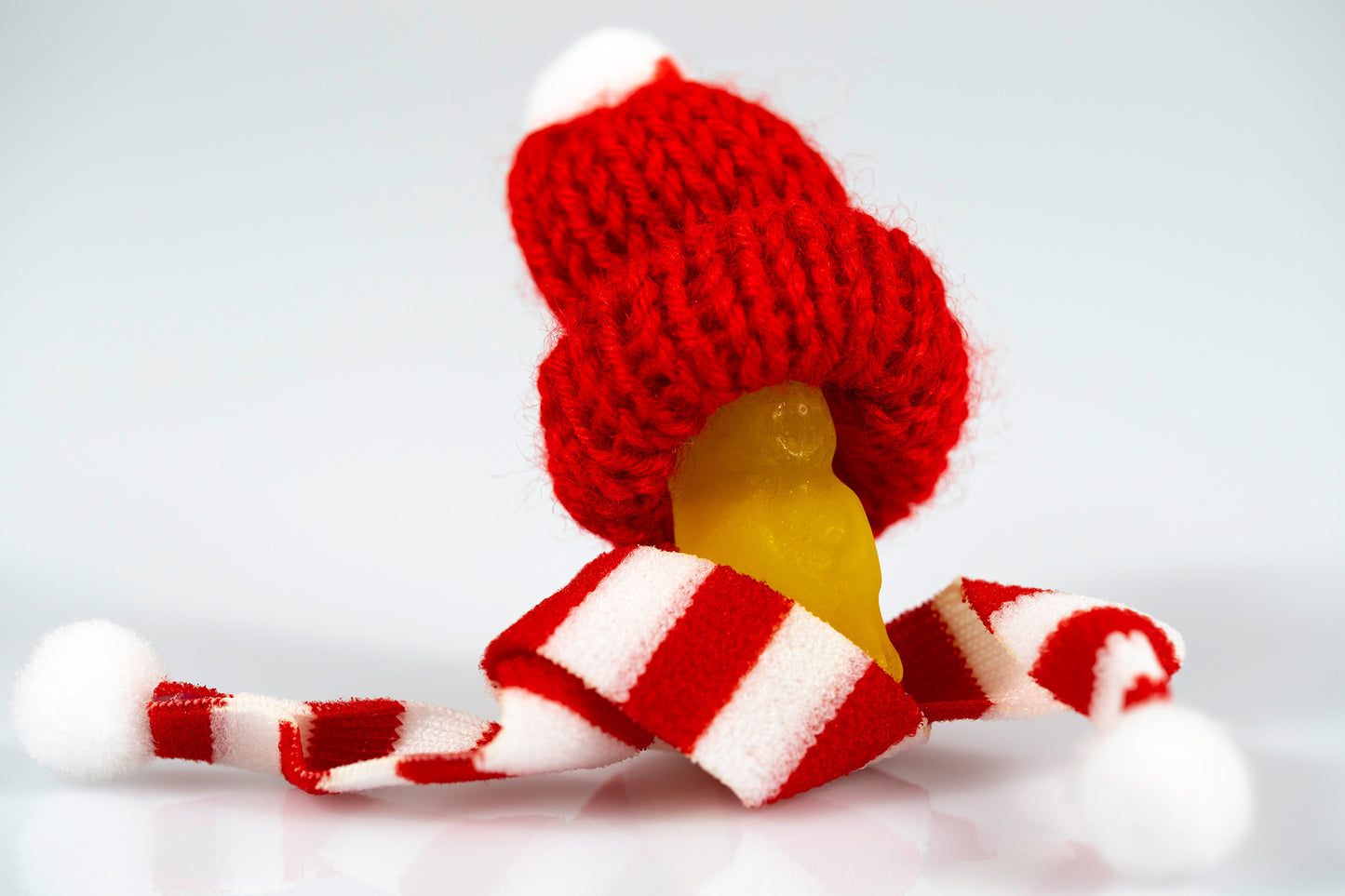 Yellow miniature figure surrounded by oversized red-and-white Santa hat and scarf on a reflective white surface.