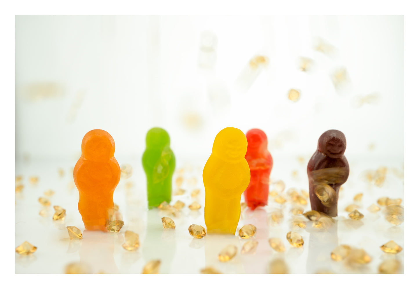 All that Glitters 2871v2 - 104x74cm, colourful jelly baby figurines in orange, green, yellow, red, and purple standing upright among scattered golden crystal fragments on a glossy white surface; playful, pop-art style fine art print by Raymond Mather, ART1.