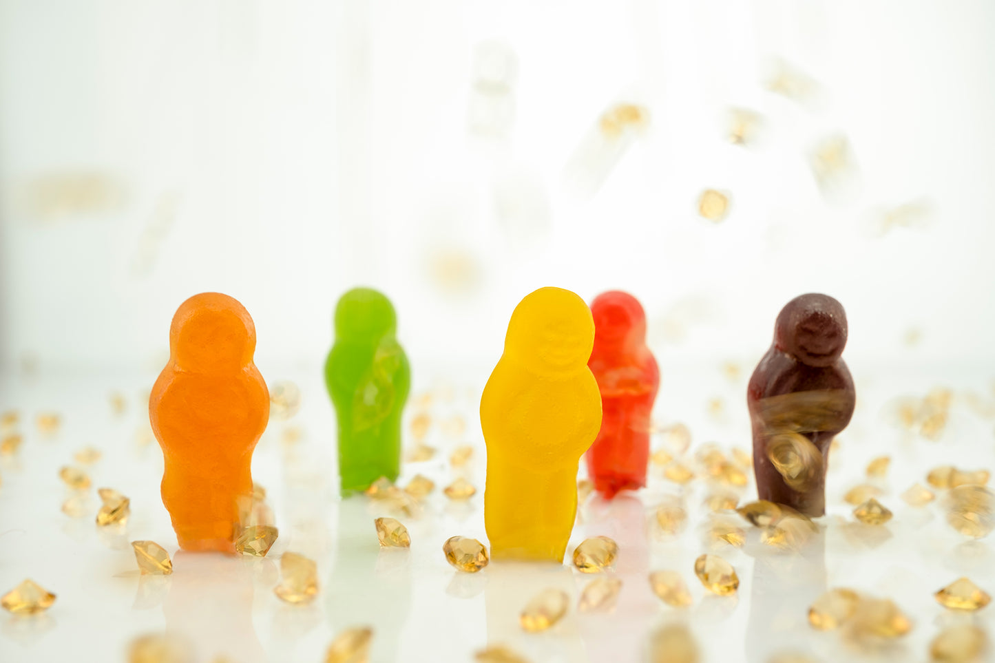 All that Glitters 2871v2 - 90x60cm, colourful jelly baby figurines in orange, green, yellow, red, and purple standing upright among scattered golden crystal fragments on a glossy white surface; playful, pop-art style fine art print by Raymond Mather, ART1.