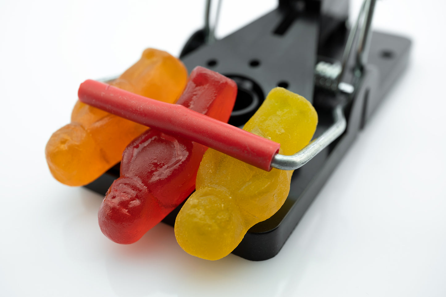 Caught in a Trap 3027 - 90x60cm, fine art photograph of three gummy jelly figures caught beneath the bar of a black metal trap, captured with precision and pop-art wit by Raymond Mather, ART1.
