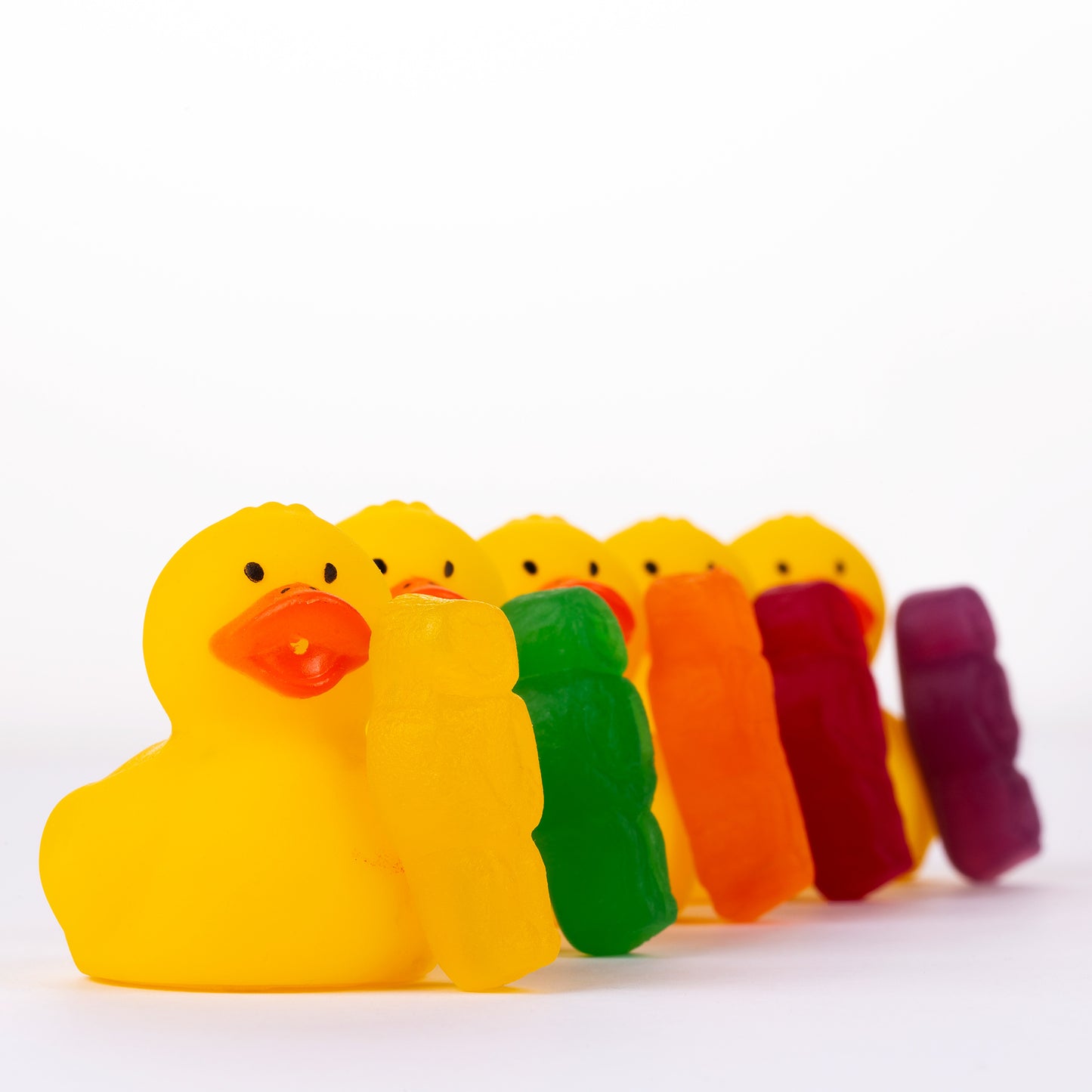 Ducks in a Row 1519 - 90x90cm, vibrant jelly baby figures leaning against a line of yellow rubber duckies, forming a rainbow procession on a white background, fine art photography by Raymond Mather, ART1.