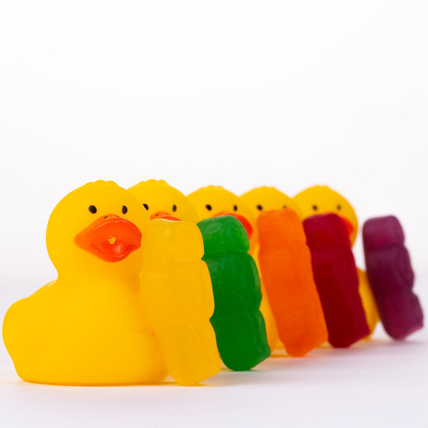 Ducks in a Row 1519 - Close Detail View, vibrant jelly baby figures leaning against a line of yellow rubber duckies, forming a rainbow procession on a white background, fine art photography by Raymond Mather, ART1.