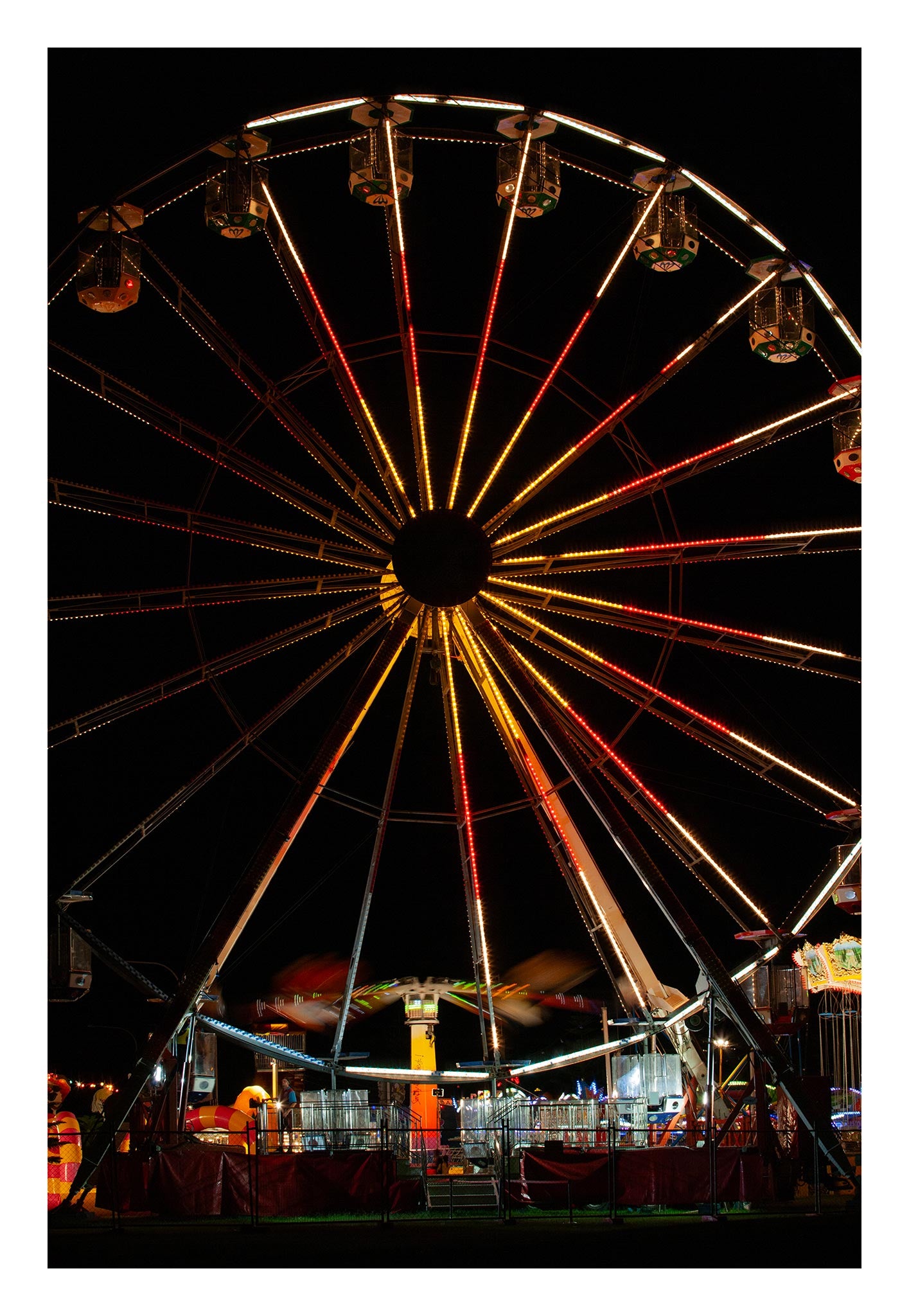 Fairground Carnival Lights 6900065 - 74x104cm, a brightly lit Ferris wheel dominates the night sky, its radiant red, orange, and gold lights forming a dazzling starburst above the motion blur of a spinning ride below. Captured by Raymond Mather, this fine art image evokes the joyful energy and nostalgic charm of a bustling carnival.