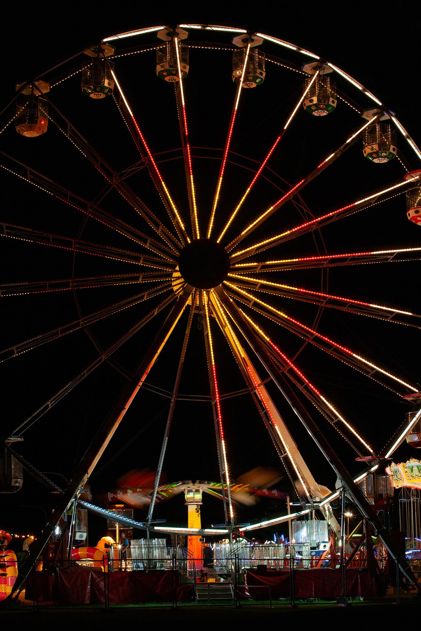 Fairground Carnival Lights 6900065 - 60x90cm, a brightly lit Ferris wheel dominates the night sky, its radiant red, orange, and gold lights forming a dazzling starburst above the motion blur of a spinning ride below. Captured by Raymond Mather, this fine art image evokes the joyful energy and nostalgic charm of a bustling carnival.