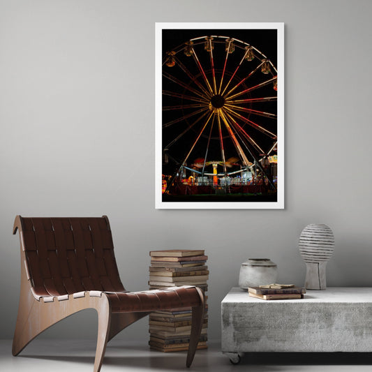 Fairground Carnival Lights 6900065 - Room View, a brightly lit Ferris wheel dominates the night sky, its radiant red, orange, and gold lights forming a dazzling starburst above the motion blur of a spinning ride below. Captured by Raymond Mather, this fine art image evokes the joyful energy and nostalgic charm of a bustling carnival
