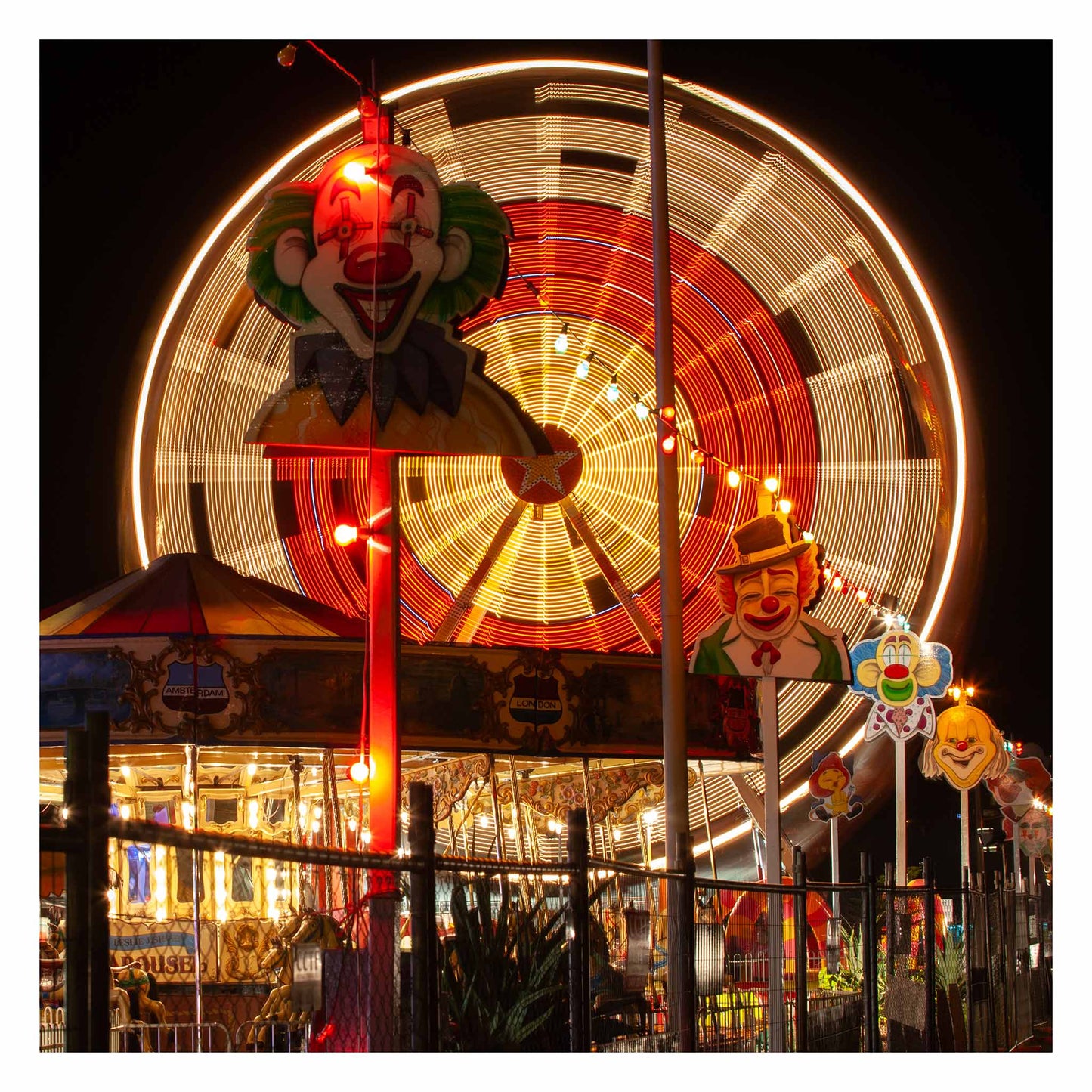 Fairground Carnival Lights 6900090 - 104x104cm, a vivid scene of a spinning Ferris wheel captured in long exposure behind a row of illuminated clown faces and a carousel. Glowing with reds and golds, the image by Raymond Mather evokes the spirited atmosphere of carnival nights, filled with nostalgia, laughter, and motion.