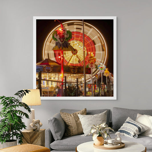 Fairground Carnival Lights 6900090 - Room View, a vivid scene of a spinning Ferris wheel captured in long exposure behind a row of illuminated clown faces and a carousel. Glowing with reds and golds, the image by Raymond Mather evokes the spirited atmosphere of carnival nights, filled with nostalgia, laughter, and motion.