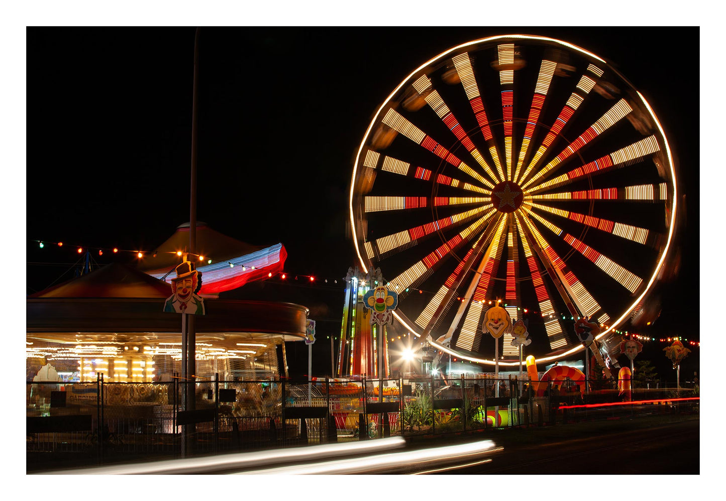 Fairground Carnival Lights 6900103 - 104x74cm, a nighttime carnival scene with a glowing Ferris wheel in spinning motion, colourful string lights, carousel, and clown signs in the foreground. A long exposure adds movement to the entire image, capturing the vibrant pulse of fairground life through Raymond Mather’s lens.