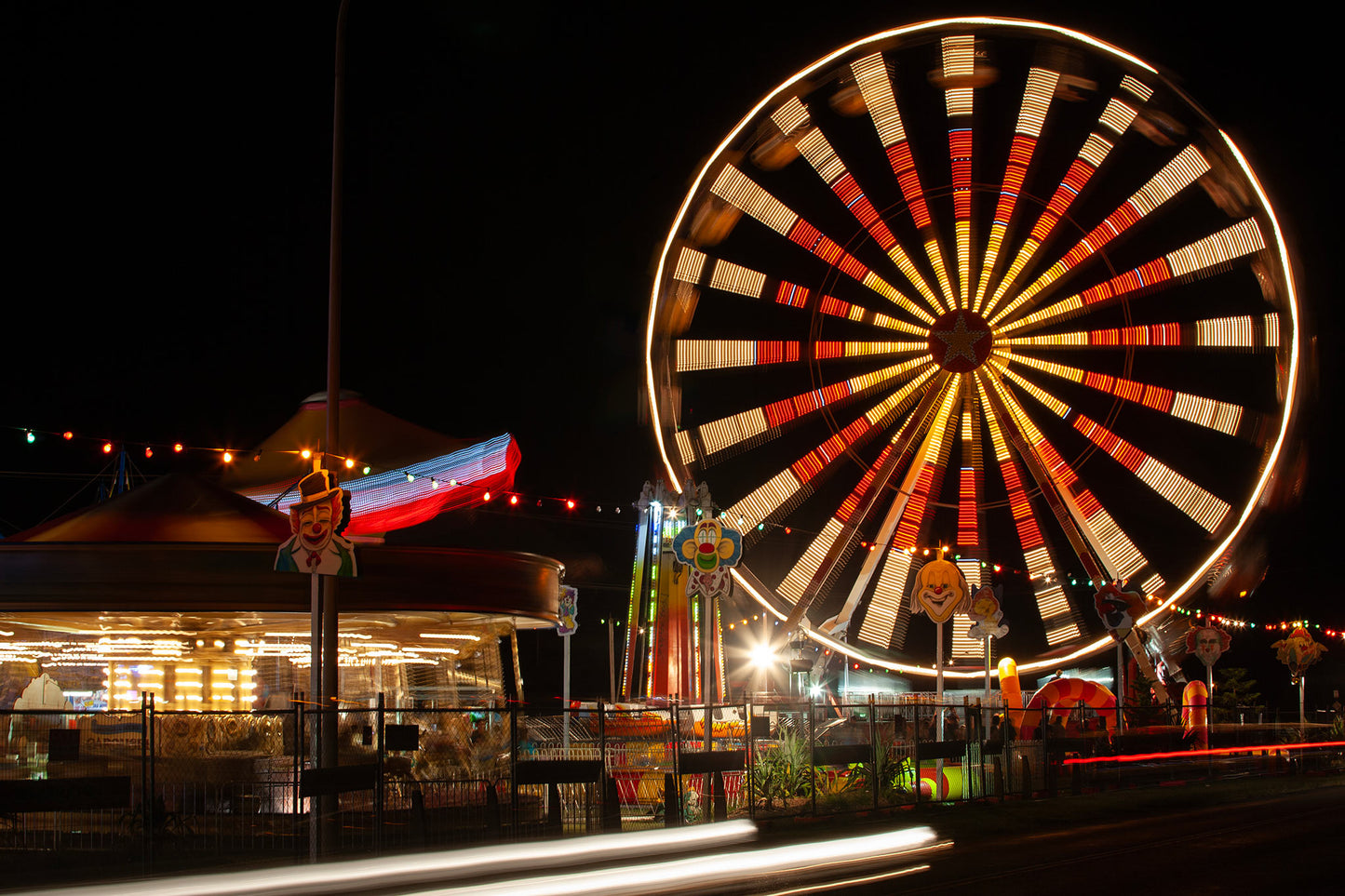 Fairground Carnival Lights 6900103 - 90x60cm, a nighttime carnival scene with a glowing Ferris wheel in spinning motion, colourful string lights, carousel, and clown signs in the foreground. A long exposure adds movement to the entire image, capturing the vibrant pulse of fairground life through Raymond Mather’s lens.