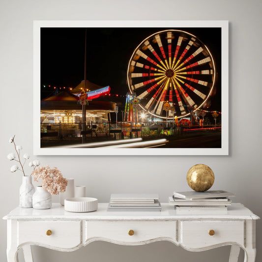 Fairground Carnival Lights 6900103 - Room View, a nighttime carnival scene with a glowing Ferris wheel in spinning motion, colourful string lights, carousel, and clown signs in the foreground. A long exposure adds movement to the entire image, capturing the vibrant pulse of fairground life through Raymond Mather’s lens.