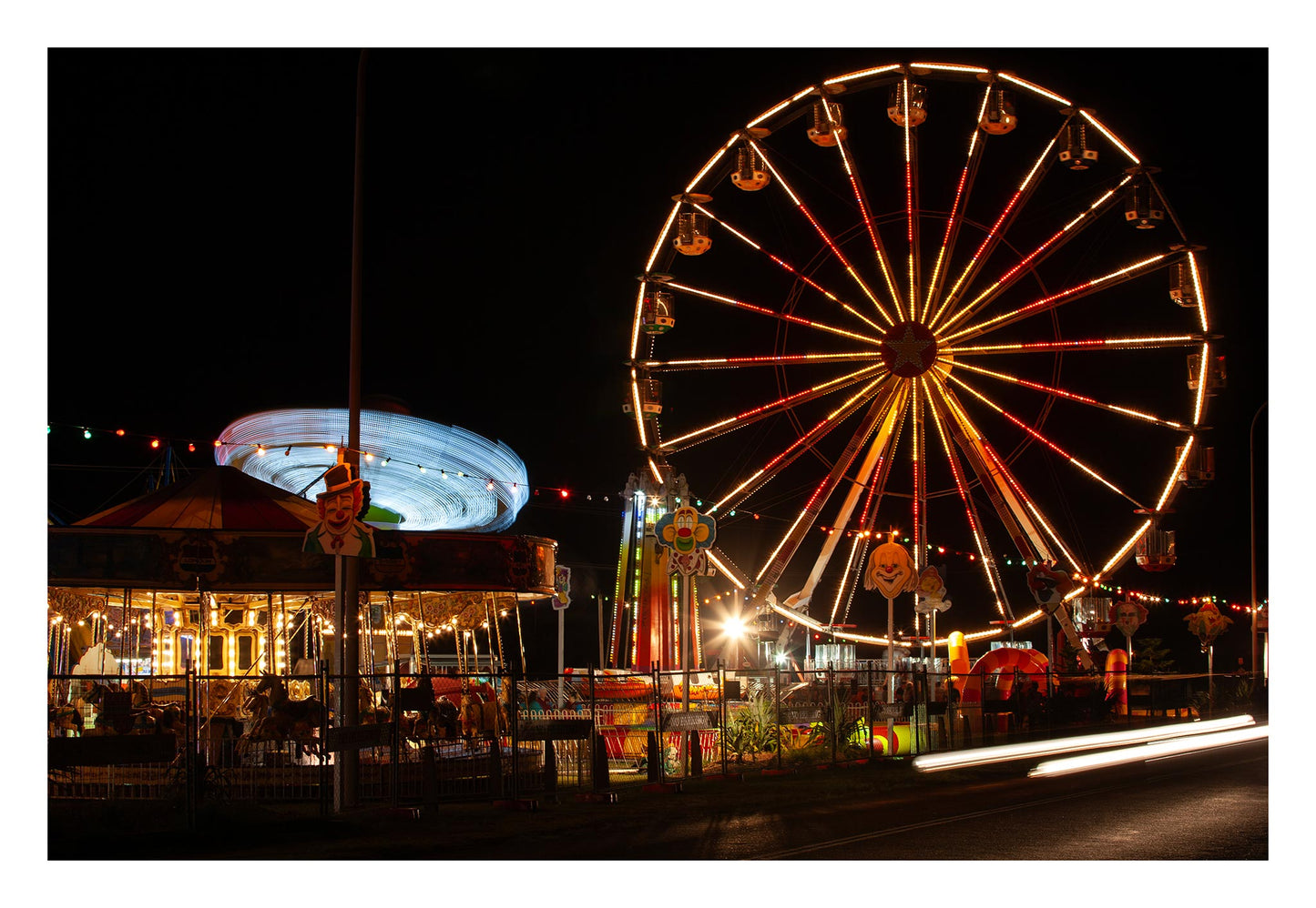 Fairground Carnival Lights 6900108 - 104x74cm, a long exposure nighttime image capturing a glowing Ferris wheel, a spinning carousel in streaks of blue light, and vibrant fairground décor including clown signs and string lights. The photograph by Raymond Mather captures the immersive energy and joy of carnival nights.