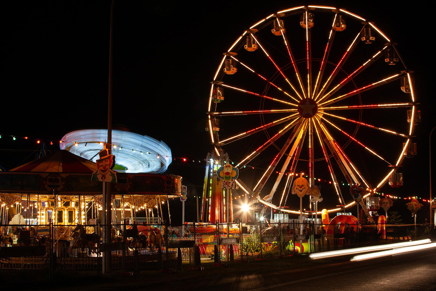 Fairground Carnival Lights 6900108 - 90x60cm, a long exposure nighttime image capturing a glowing Ferris wheel, a spinning carousel in streaks of blue light, and vibrant fairground décor including clown signs and string lights. The photograph by Raymond Mather captures the immersive energy and joy of carnival nights.