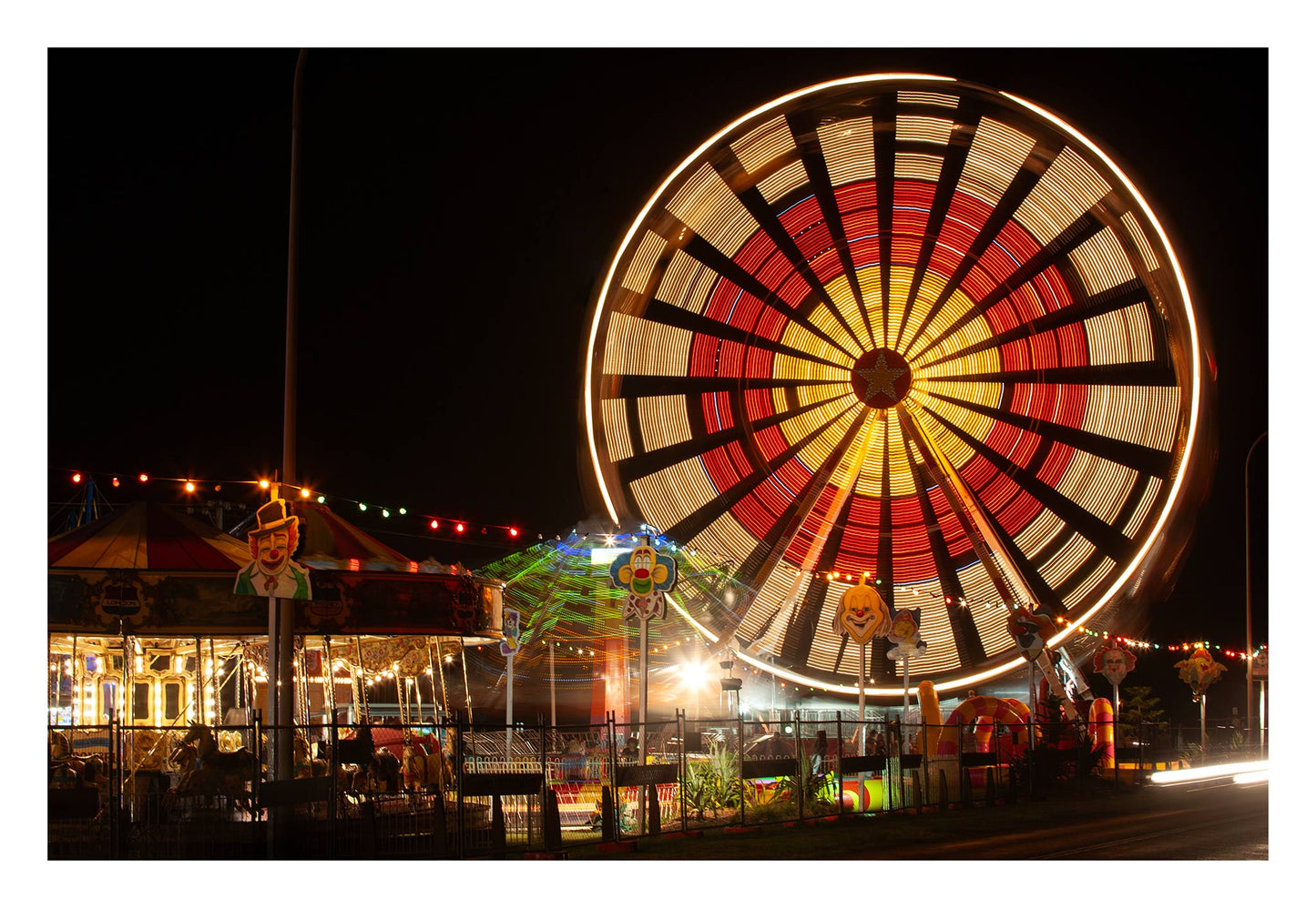 Fairground Carnival Lights 6900129 - 104x74cm, a vibrant Ferris wheel spins in glowing red, yellow, and white light, creating a radiant circular pattern. Below it, a carousel and other rides glow with multicoloured lights and vintage clown decorations. Photographed by Raymond Mather, this image evokes the sensory thrill and nostalgia of carnival nights.