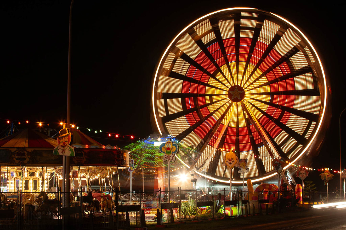 Fairground Carnival Lights 6900129 - 90x60cm, a vibrant Ferris wheel spins in glowing red, yellow, and white light, creating a radiant circular pattern. Below it, a carousel and other rides glow with multicoloured lights and vintage clown decorations. Photographed by Raymond Mather, this image evokes the sensory thrill and nostalgia of carnival nights.