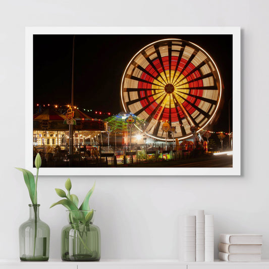 Fairground Carnival Lights 6900129 - Room View, a vibrant Ferris wheel spins in glowing red, yellow, and white light, creating a radiant circular pattern. Below it, a carousel and other rides glow with multicoloured lights and vintage clown decorations. Photographed by Raymond Mather, this image evokes the sensory thrill and nostalgia of carnival nights.