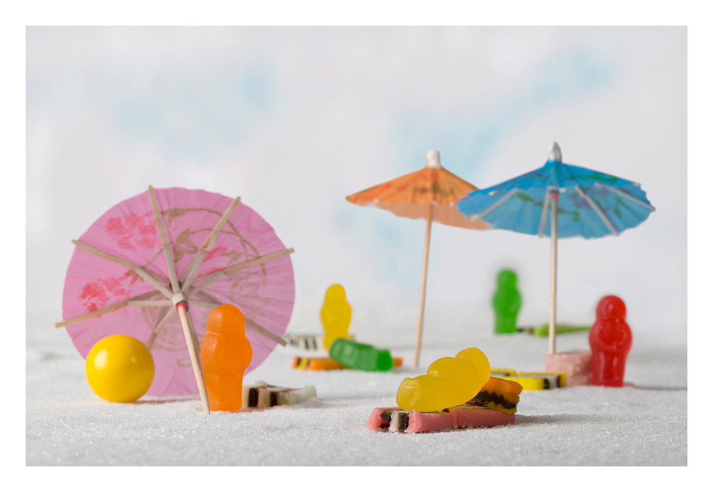 Life’s a Beach 1395 - 104x74cm, colourful jelly baby figures staged on a sugar-sand beach with cocktail umbrellas, licorice towels, and a yellow beach ball, fine art still life by Raymond Mather, ART1.