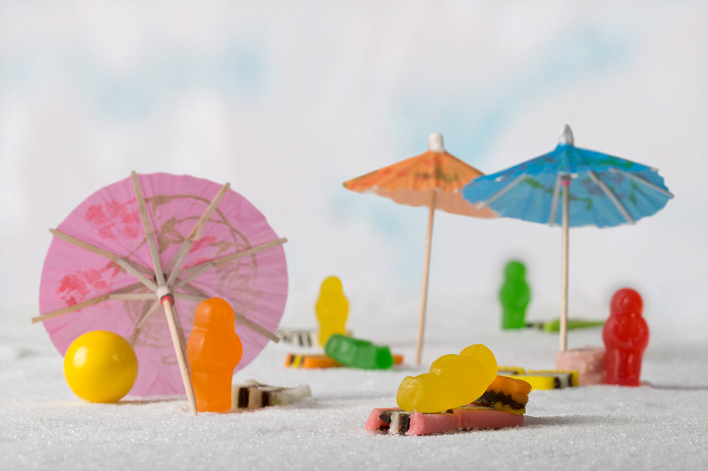 Life’s a Beach 1395 - 90x60cm, colourful jelly baby figures staged on a sugar-sand beach with cocktail umbrellas, licorice towels, and a yellow beach ball, fine art still life by Raymond Mather, ART1.