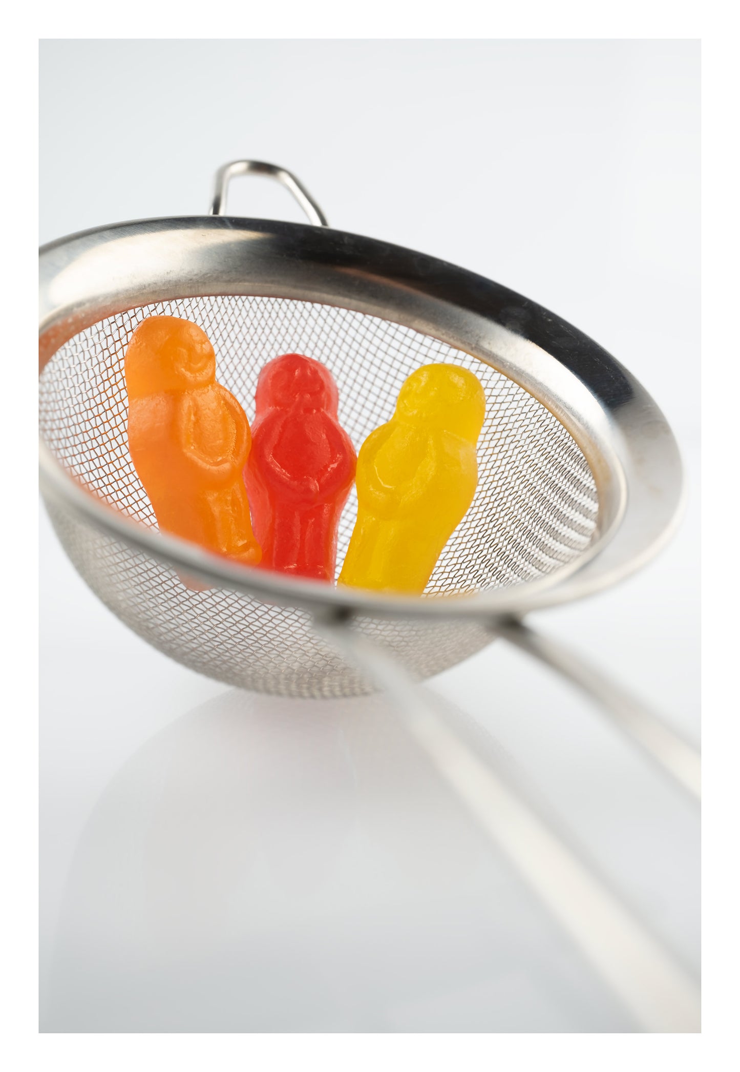 Life’s a Strain 2541v1 - 74x104cm, stylised fine art photograph featuring three vibrant jelly baby figures, orange, red, and yellow, nestled inside a silver tea strainer against a soft white background. A whimsical composition evoking themes of containment, reflection, and resilience, by Raymond Mather, ART1.
