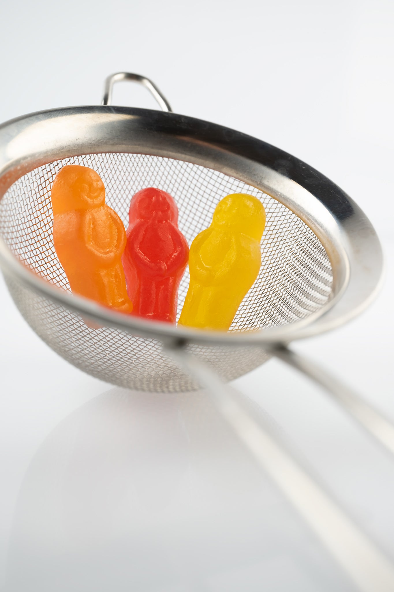 Life’s a Strain 2541v1 - 60x90cm, stylised fine art photograph featuring three vibrant jelly baby figures, orange, red, and yellow, nestled inside a silver tea strainer against a soft white background. A whimsical composition evoking themes of containment, reflection, and resilience, by Raymond Mather, ART1.