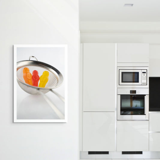 Life’s a Strain 2541v1 - Room View, stylised fine art photograph featuring three vibrant jelly baby figures, orange, red, and yellow, nestled inside a silver tea strainer against a soft white background. A whimsical composition evoking themes of containment, reflection, and resilience, by Raymond Mather, ART1.