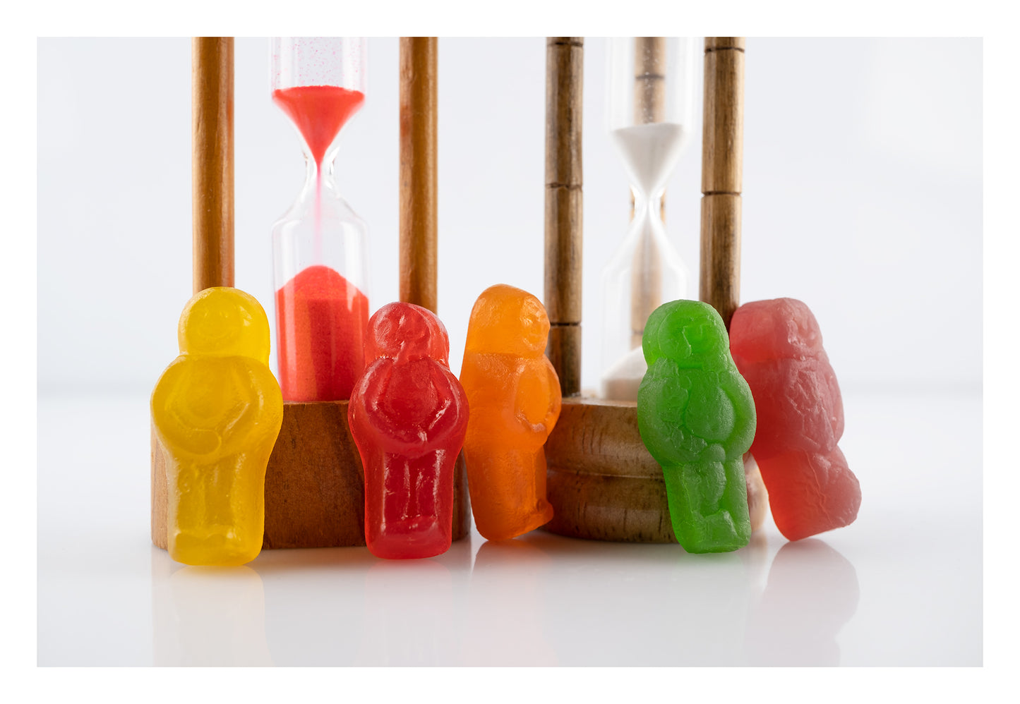 Like Sands Through the Hourglass 2683v1 - 104x74cm, whimsical still life of colourful jelly baby figures standing beside vintage sand timers with red and white sand, reflecting time, nostalgia, and childhood in a playful conceptual fine art photograph by Raymond Mather, ART1.