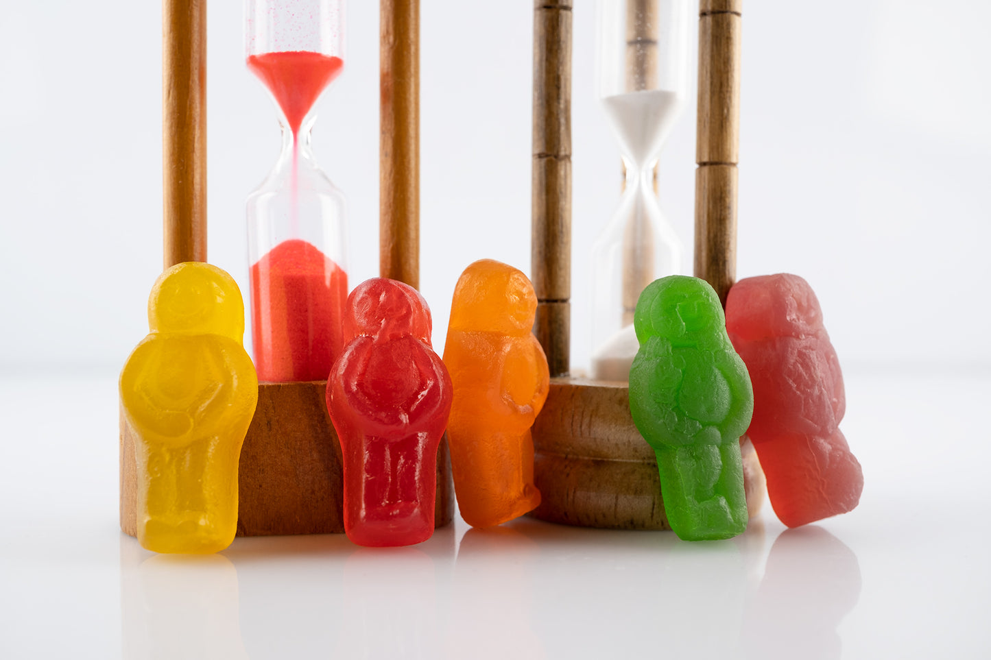 Like Sands Through the Hourglass 2678v1 - 90x60cm, whimsical still life of colourful jelly baby figures standing beside vintage sand timers with red and white sand, reflecting time, nostalgia, and childhood in a playful conceptual fine art photograph by Raymond Mather, ART1.