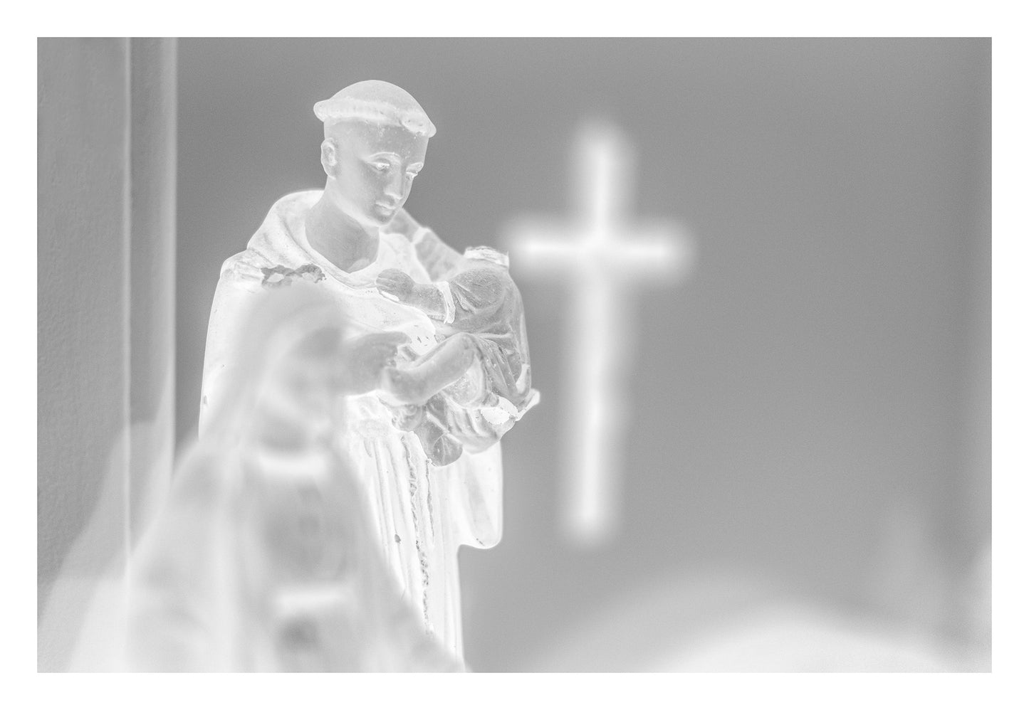 Monochrome Mix - Icons on the Mantel - 183102 - 104x74cm, Black and white image of a Saint Anthony statue holding baby Jesus, with a glowing cross in the blurred background, evoking spiritual reflection and reverent stillness.