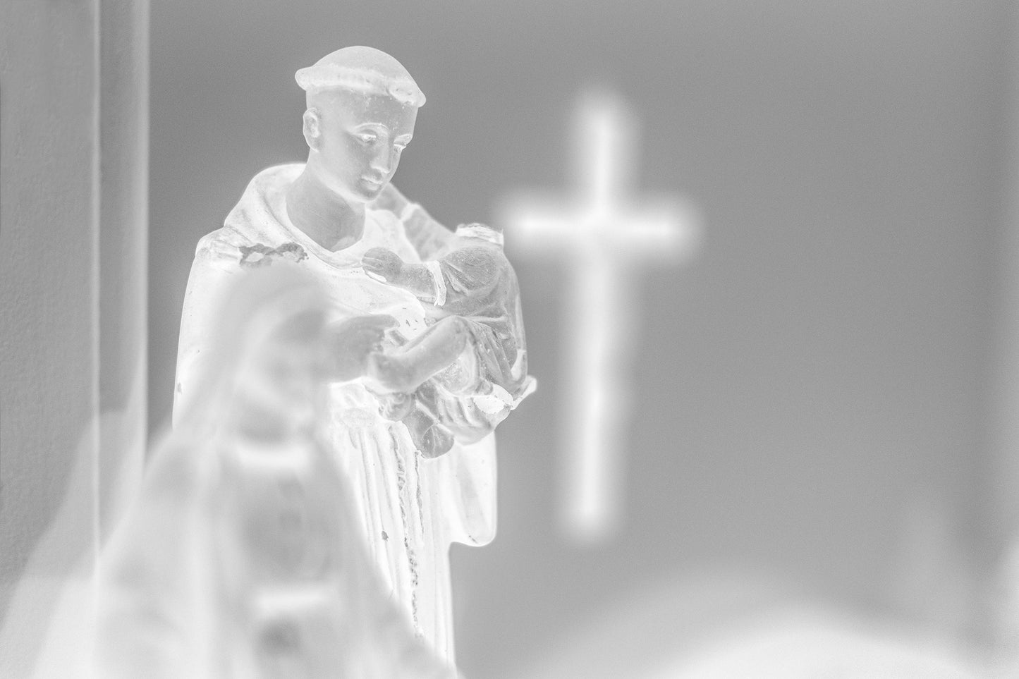 Monochrome Mix - Icons on the Mantel - 183102 - 90x60cm, Black and white image of a Saint Anthony statue holding baby Jesus, with a glowing cross in the blurred background, evoking spiritual reflection and reverent stillness.