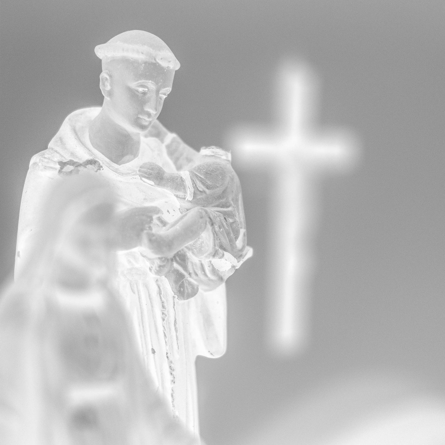 Monochrome Mix - Icons on the Mantel - 183102 - Close Detail View, Black and white image of a Saint Anthony statue holding baby Jesus, with a glowing cross in the blurred background, evoking spiritual reflection and reverent stillness.