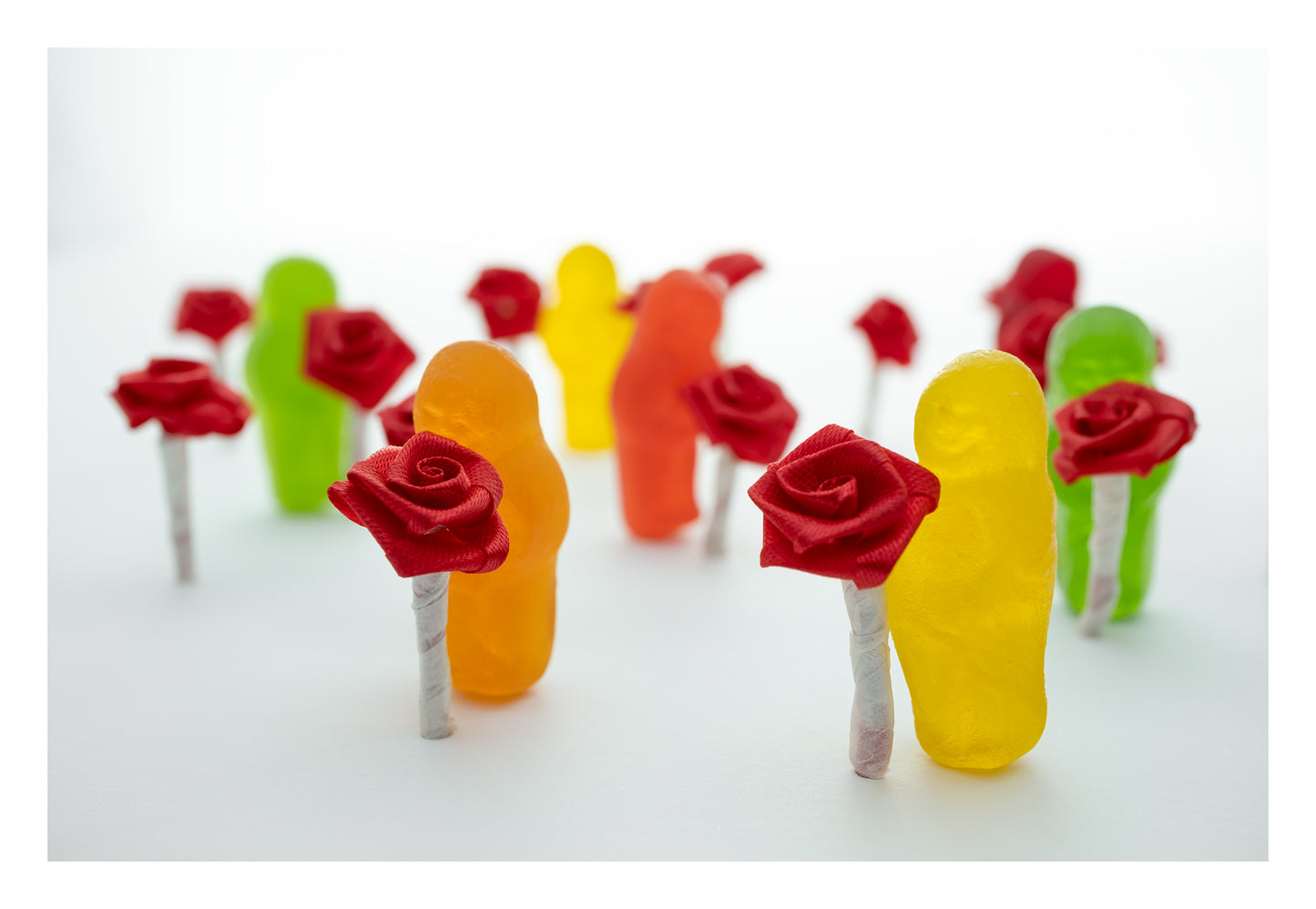 Stop and Smell the Roses 2836v1 - 104x74cm, playful fine art photograph by Raymond Mather featuring colourful jelly baby figures holding miniature red ribbon roses, arranged in a dreamlike procession on a soft white backdrop, evoking mindfulness and delight.