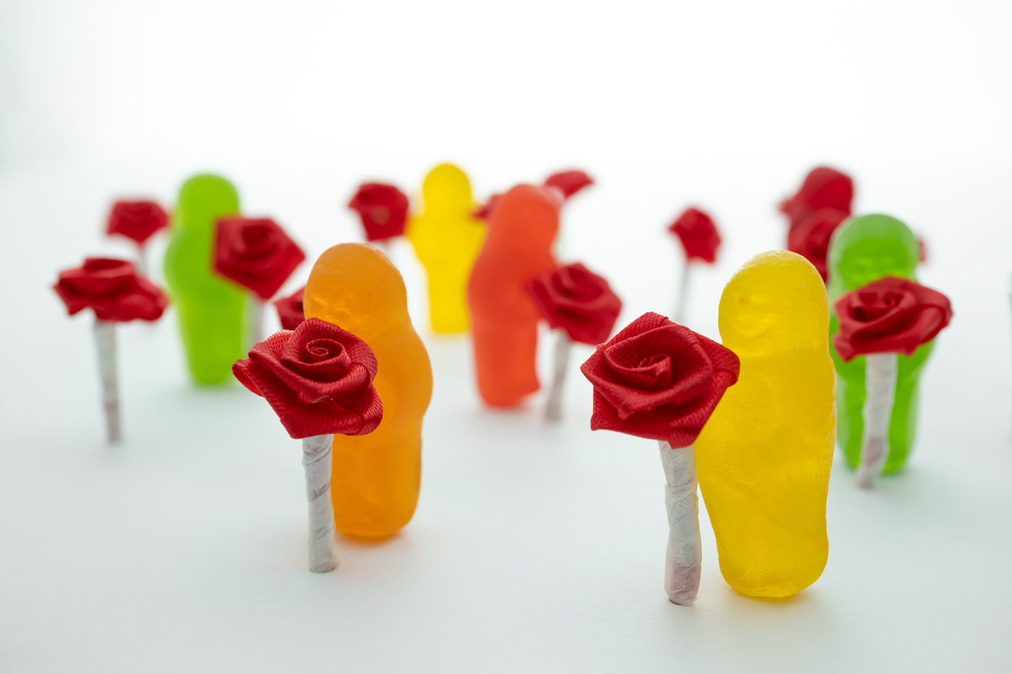 Stop and Smell the Roses 2836v1 - 90x60cm, playful fine art photograph by Raymond Mather featuring colourful jelly baby figures holding miniature red ribbon roses, arranged in a dreamlike procession on a soft white backdrop, evoking mindfulness and delight.