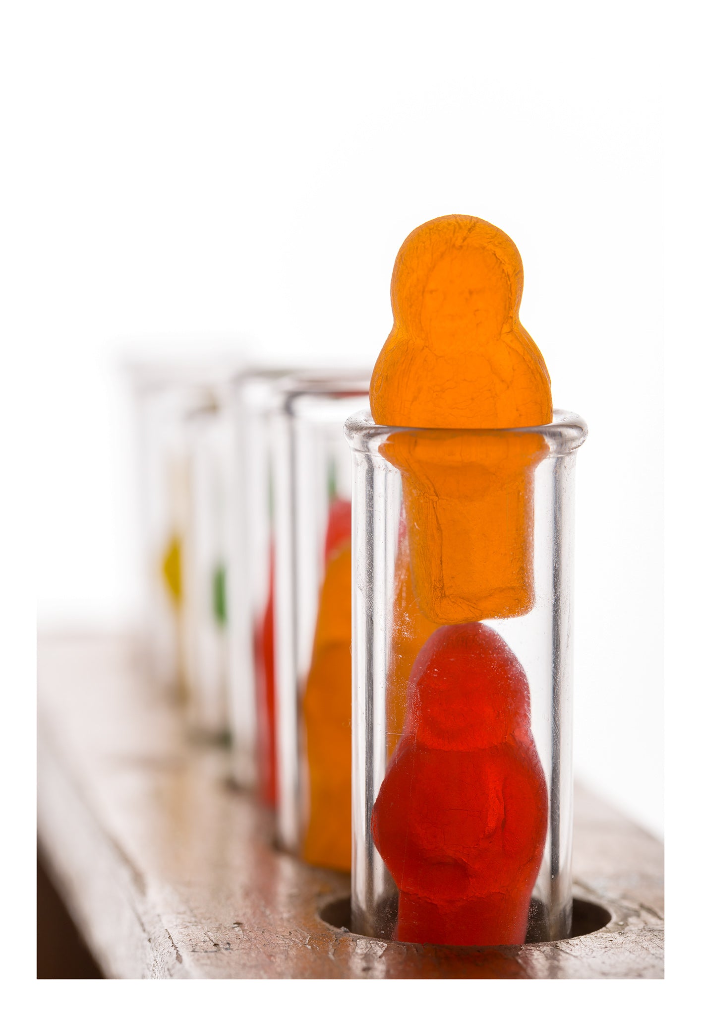 Test Tube Babies 20101 - 74x104cm, fine art photograph by Raymond Mather featuring brightly coloured jelly baby figures inside clear glass test tubes arranged in a wooden rack, a playful yet thought provoking pop art tableau exploring identity, science, and the absurdity of cliché.