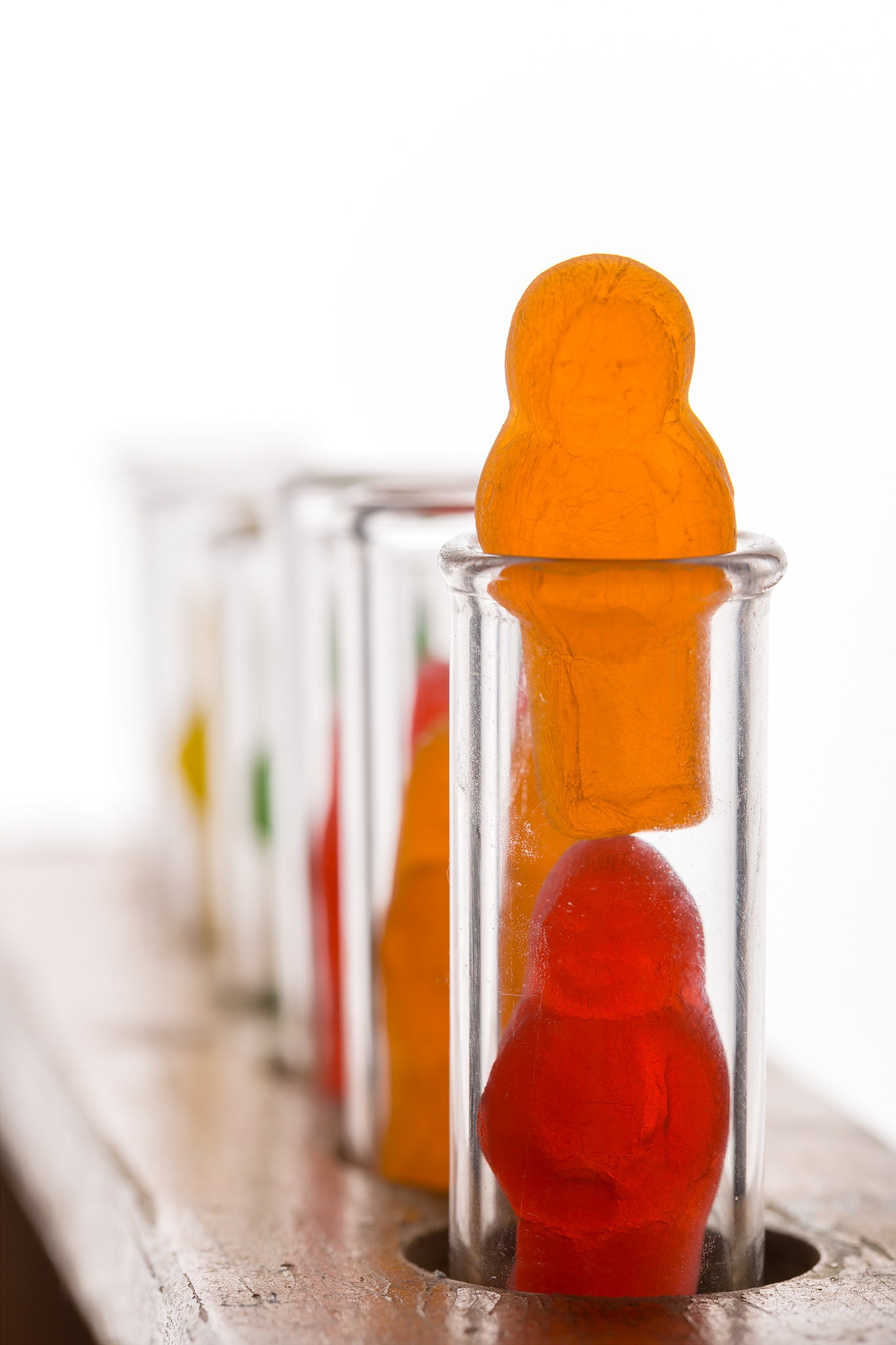 Test Tube Babies 20101 - 60x90cm, fine art photograph by Raymond Mather featuring brightly coloured jelly baby figures inside clear glass test tubes arranged in a wooden rack, a playful yet thought provoking pop art tableau exploring identity, science, and the absurdity of cliché.