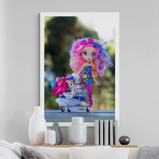 Trolley Dolly - Series No.1: 197260 - Room View, a colourful fashion doll with curly rainbow hair and a tear on her cheek stands beside an overflowing miniature shopping trolley packed with symbolic items like ribbons, fabric, and string. Set against a soft outdoor backdrop, the image explores displacement, resilience, and hidden stories.