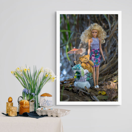 Trolley Dolly - Series No.1: 197274 - Room View, a blonde fashion doll stands in natural surroundings beside a miniature shopping trolley filled with symbolic items, including a teddy bear, paper parasol, plastic bag, and folded cloth. The image explores homelessness, innocence, and feminine resilience.