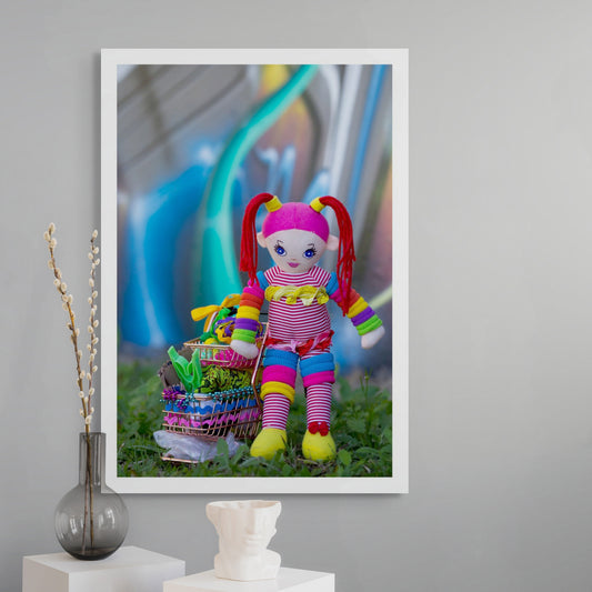 Trolley Dolly - Series No.1: 197302 - Room View, a handmade rainbow coloured rag doll stands beside a mini shopping trolley filled with ribbons, beads, fabric, and found objects. Set on grass against a vivid urban graffiti wall, the image explores homelessness, survival, and innocence.