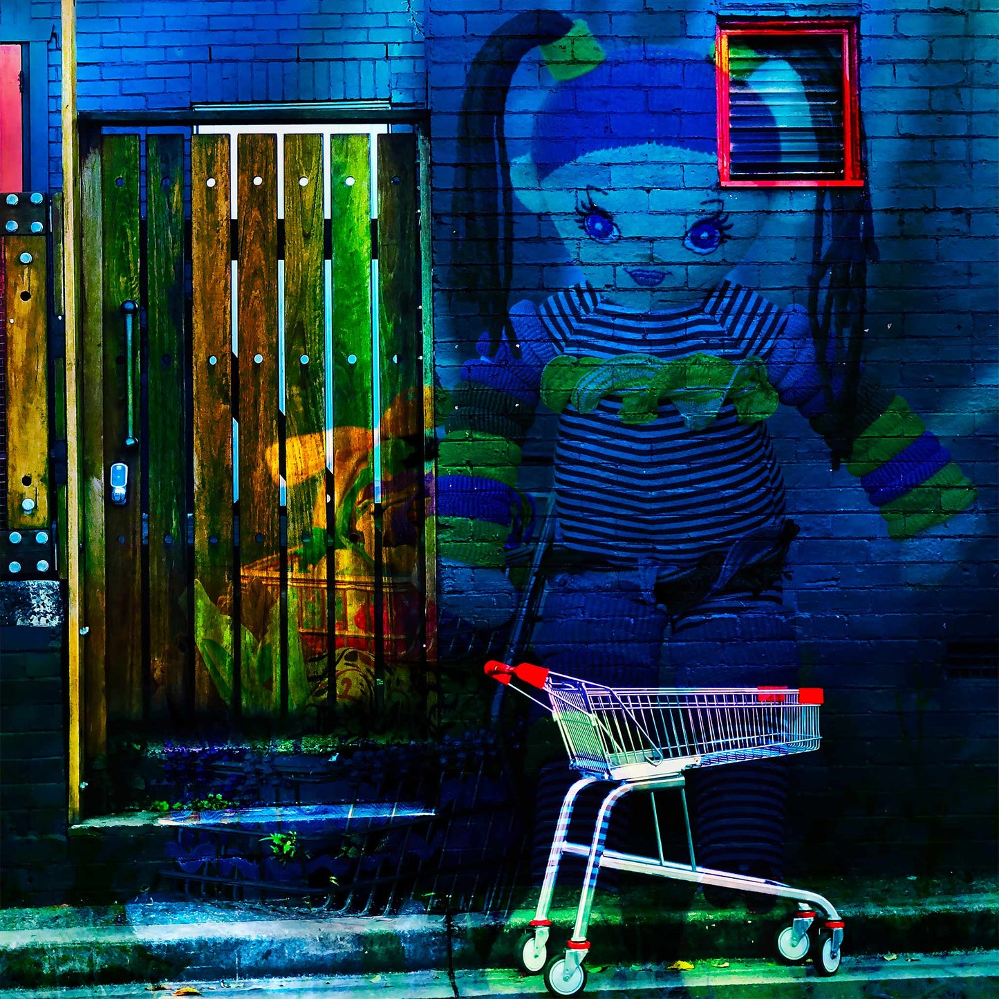 Trolley Dolly - Series No.2: 19E2255v14 - 60x60cm, a knitted doll with striped clothing and ponytails appears against a dark blue brick wall. A red-handled shopping trolley sits below, and a wooden door to the left glows with market textures and shadow. The surreal image explores homelessness and emotional vulnerability.