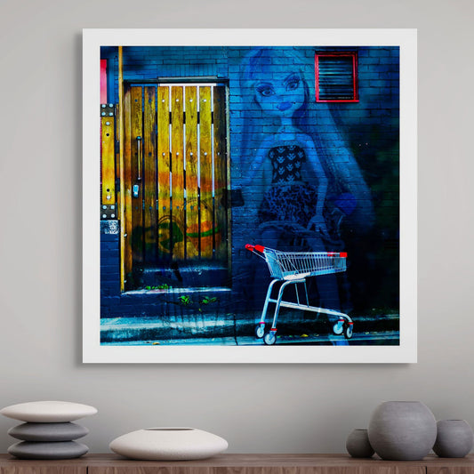 Trolley Dolly - Series No.2: 19E2255 - Room View, a fine art photograph featuring a whimsical street mural of a stylised female doll figure painted on a dark blue brick wall beside a rustic wooden door, with a red-and-silver shopping trolley in the foreground. The vibrant image contrasts playful pop culture with gritty urban textures.