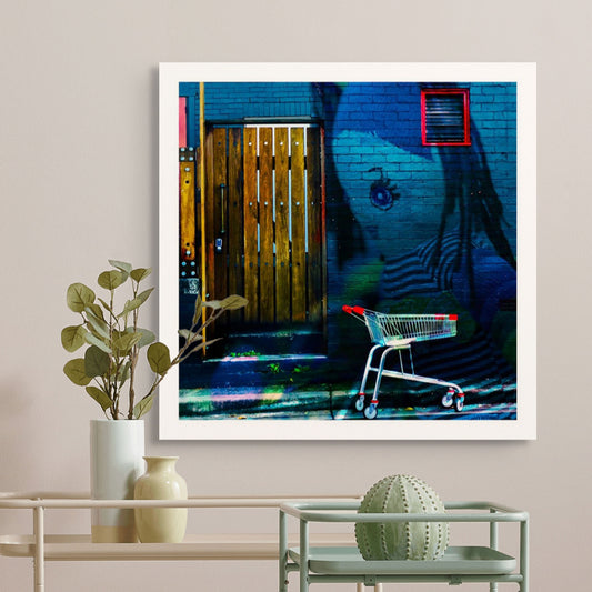 Trolley Dolly - Series No.2: 19E2255v1 - Room View, a fine art photograph featuring a large painted eye and striped figure of a girl on a blue brick wall, beside a rustic timber door and a red and silver shopping trolley on a weathered street. The scene captures themes of invisibility, childhood, and urban displacement.