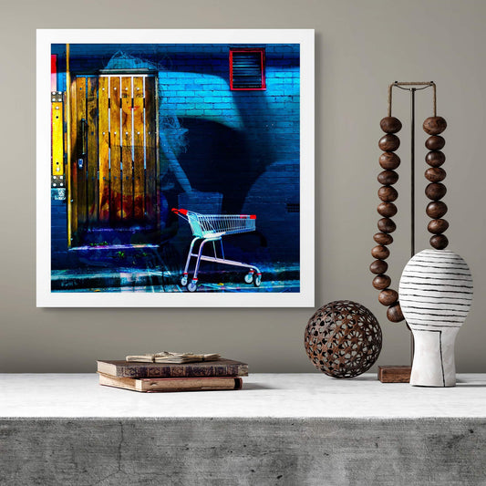 Trolley Dolly - Series No.2: 19E2255v11 - Room View, a translucent doll like figure appears on a wooden door, her face softly illuminated amid rich textures. A red handled shopping trolley sits on the pavement below. Deep shadows and a moody blue brick wall complete the scene, evoking themes of homelessness, invisibility, and survival.