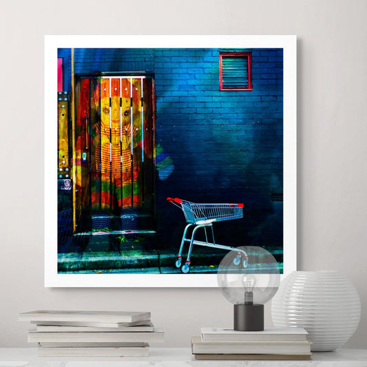 Trolley Dolly - Series No.2: 19E2255v13 - Room View, a colourful knitted doll appears embedded in a wooden door, her red, green, and striped outfit vivid against a moody blue brick wall. An empty red handled shopping trolley sits at the kerbside. The composition symbolises childhood, homelessness, and the silent resilience of unseen lives.