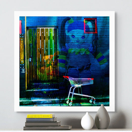 Trolley Dolly - Series No.2: 19E2255v14 - Room View, a knitted doll with striped clothing and ponytails appears against a dark blue brick wall. A red-handled shopping trolley sits below, and a wooden door to the left glows with market textures and shadow. The surreal image explores homelessness and emotional vulnerability.