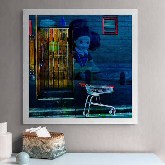 Trolley Dolly - Series No.2: 19E2255v16 - Room View, a dark haired rag doll with outstretched limbs is layered onto a blue brick wall beside a red handled shopping trolley. A glowing wooden door sits to the left, overlaid with fruit textures. The composition explores homelessness, emotional fatigue, and invisibility.