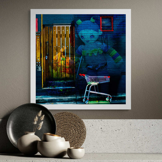 Trolley Dolly - Series No.2: 9255v3 - Room View, a surreal urban street scene featuring a large doll overlaid against a dark brick wall, her striped outfit and outstretched arms glowing in soft hues. In the foreground, a shopping trolley with red handles is parked on the footpath. A textured wooden door and a red trimmed vent hint at hidden stories. The layered composition conveys a dreamlike tension between innocence and abandonment, reflecting on homelessness and social neglect.