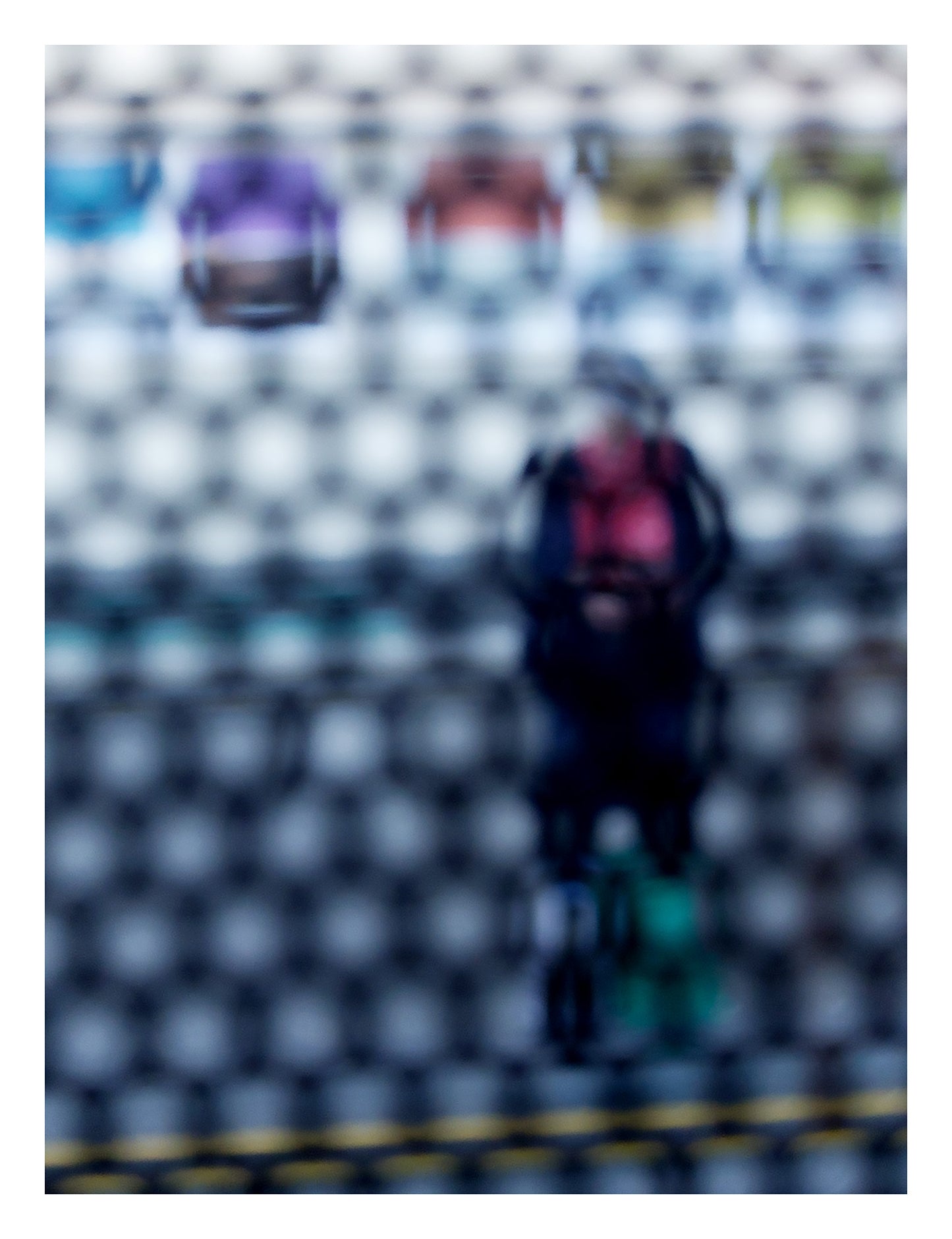 Urban Commute - Series No.2: 9153 - 81.5x104cm, fine art photograph by Raymond Mather showing a blurred figure in red and navy with a backpack and helmet, standing before a row of colourful train seats. Seen through a circular grid, the image evokes themes of pause, movement, and visual abstraction.