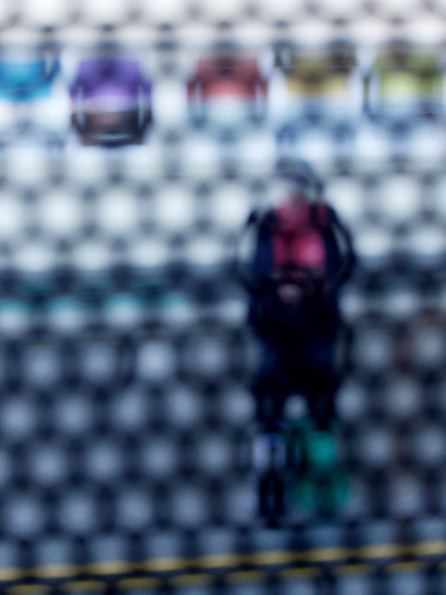 Urban Commute - Series No.2: 9153 - 67.5x90cm, fine art photograph by Raymond Mather showing a blurred figure in red and navy with a backpack and helmet, standing before a row of colourful train seats. Seen through a circular grid, the image evokes themes of pause, movement, and visual abstraction.